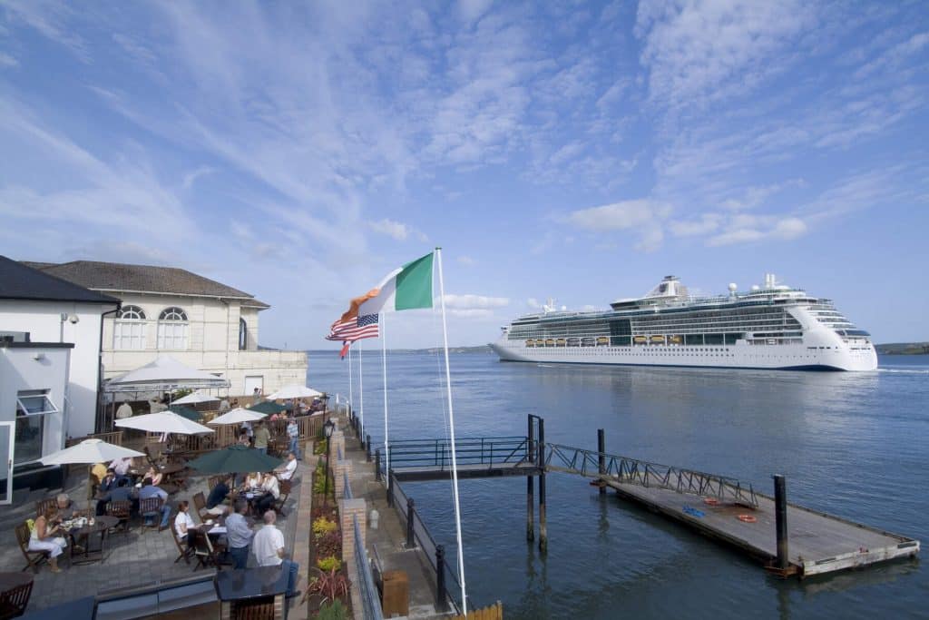 A wide-angle scenic view of a waterfront terrace with people sitting at tables under umbrellas, overlooking a body of water where a large white cruise ship is passing under a bright, clear sky.