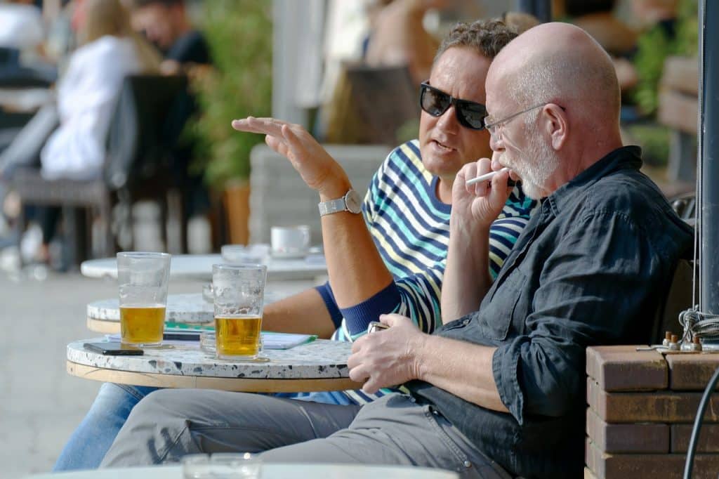 Two men sitting at an outdoor table with pints of beer, engaged in deep conversation. One man in a striped shirt gestures with his hand while the other listens intently.