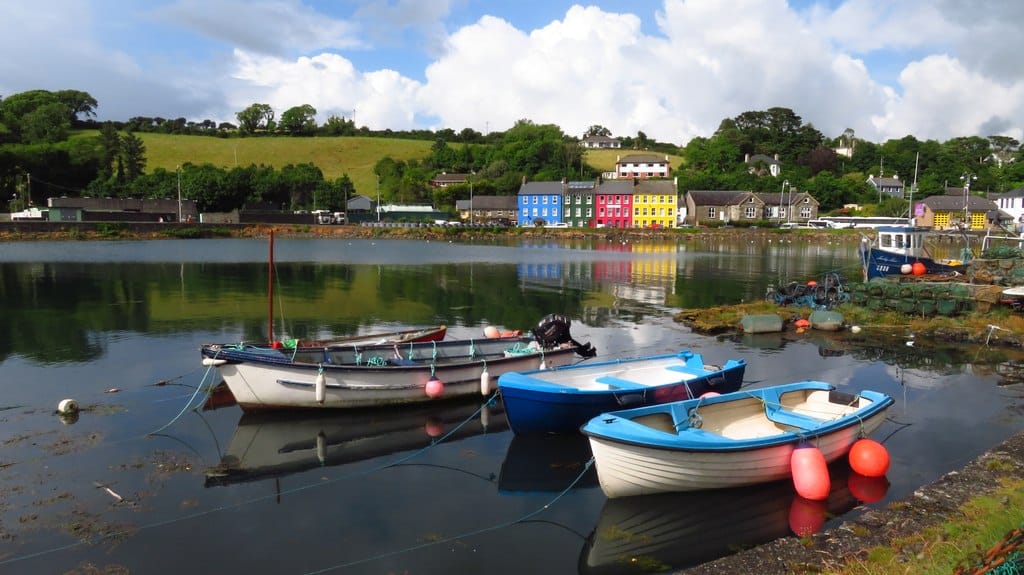 Three empty boats sit on the water in Bantry Harbour. Colourful houses and green hills make up the background.