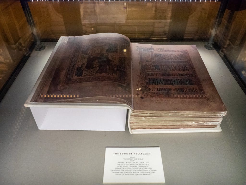 An ancient illuminated manuscript, the Book of Kells, lies open inside a glass display case with an informational placard in front.