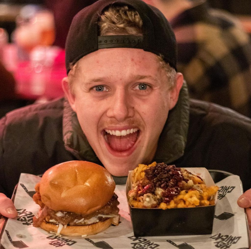 A cheerful man smiles warmly at the camera while holding his food, showcasing the vibrant culinary scene of Trademarket Belfast, with a sense of joy and satisfaction evident in his expression.