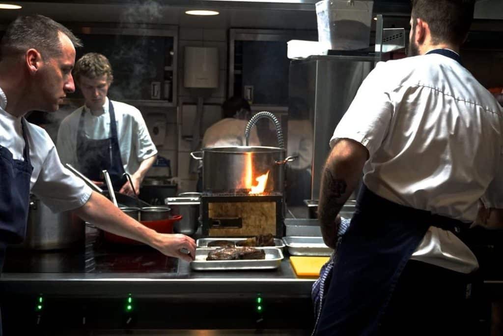 A group of chefs at Ox Restaurant in Belfast, passionately preparing a steak with precision and skill, showcasing the culinary excellence of one of Belfast's must-visit foodie destinations.