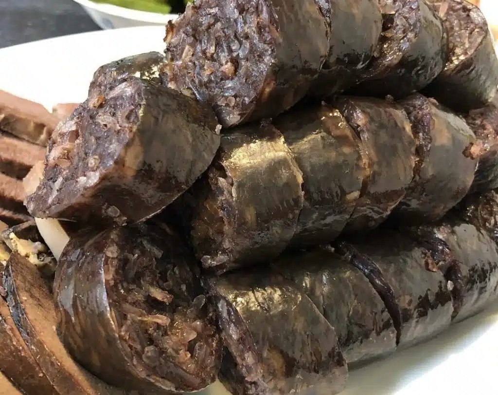 A stack of dark, sliced drisheen blood sausages served on a white plate with a side of greens.