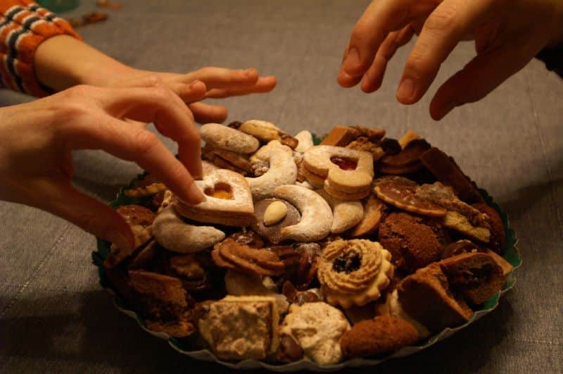 A close-up shot of a large green plate overflowing with a variety of festive biscuits and cookies, with several hands reaching toward the pile, capturing the social tension of who will take the final treats.