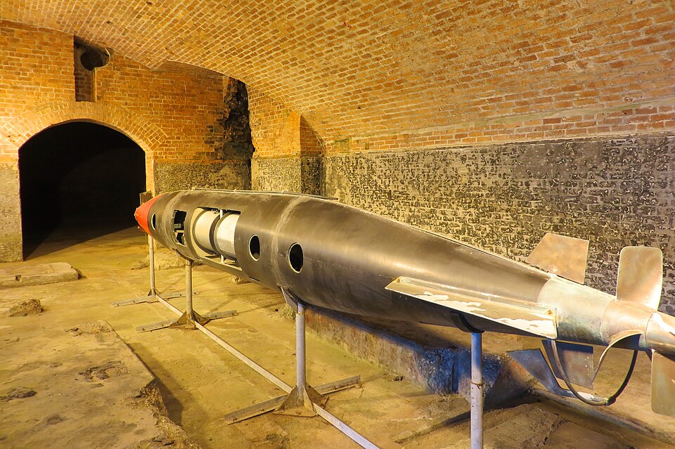 A long, black Brennan torpedo with a red tip and rear propellers displayed on metal stands inside a brick-vaulted underground bunker or fort.