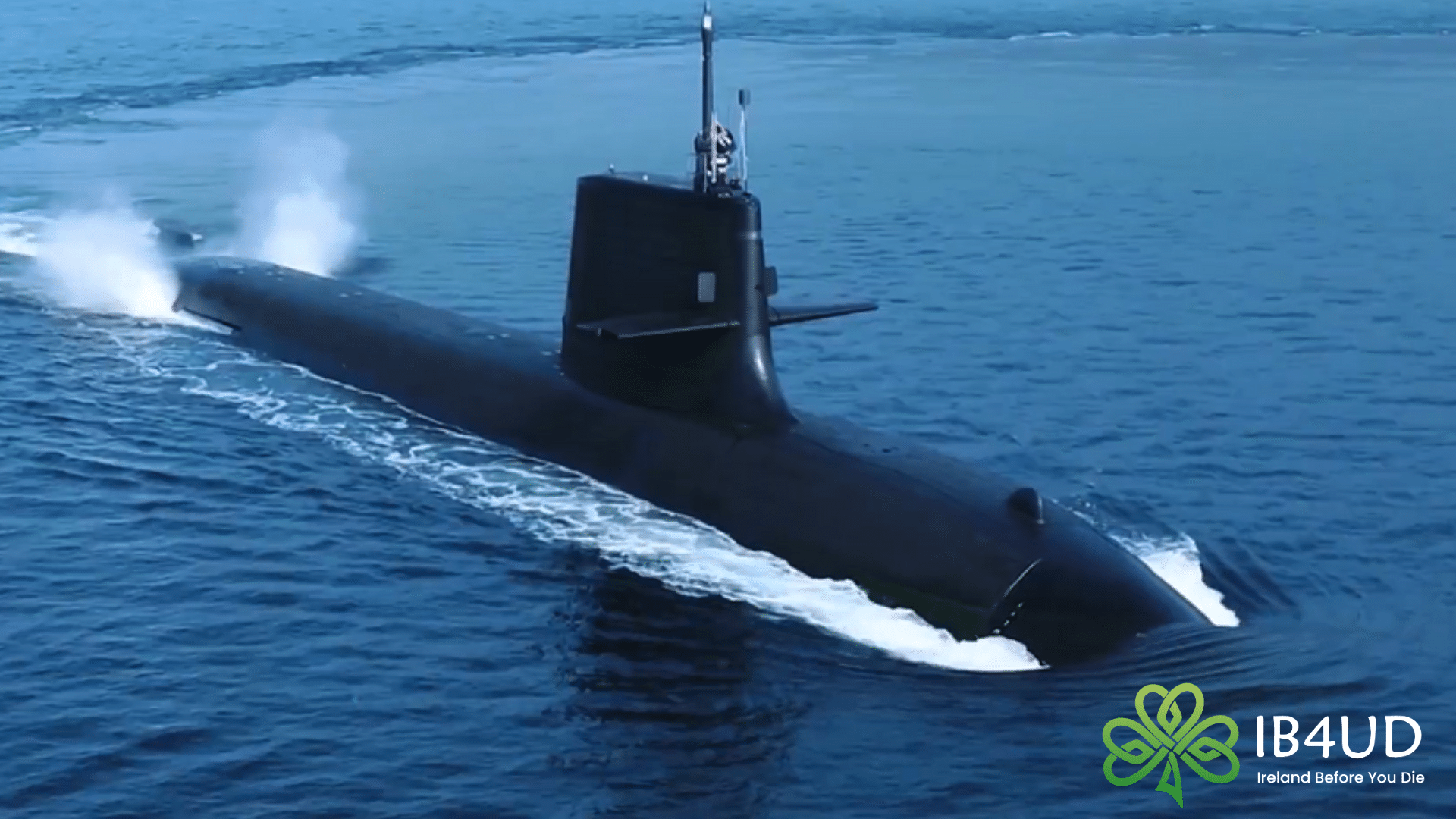 An aerial view of a black modern submarine cruising on the surface of deep blue water, leaving a white wake and spray behind it.