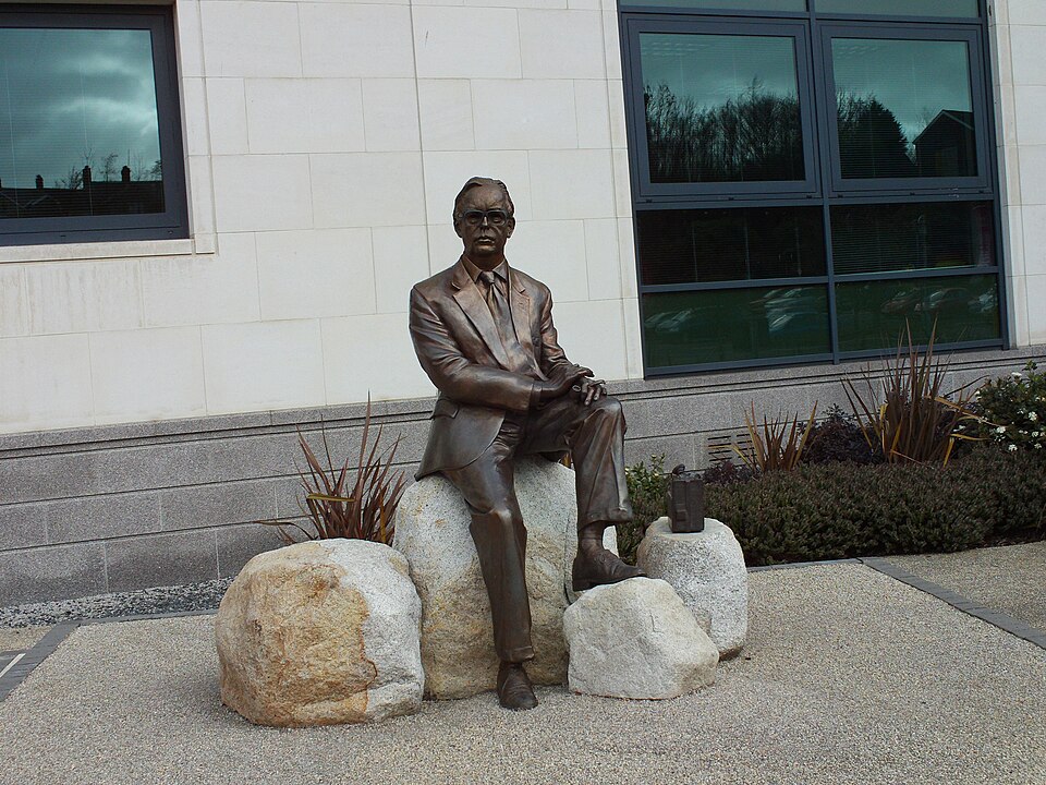 A bronze statue of Professor Frank Pantridge, the inventor of the portable defibrillator, sitting on a large rock outside a modern building.