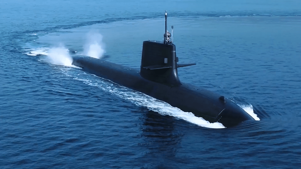 An aerial view of a black modern submarine cruising on the surface of deep blue water, leaving a white wake and spray behind it.
