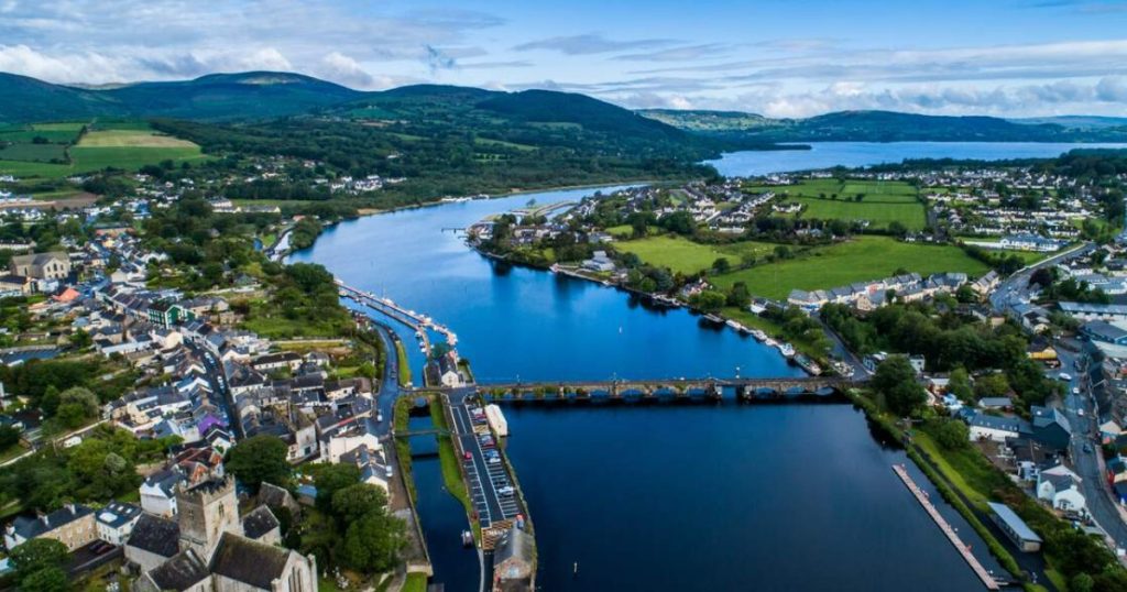 An aerial view of the twin towns of Ballina and Killaloe, connected by a historic stone bridge over the River Shannon, surrounded by rolling green hills and Lough Derg.