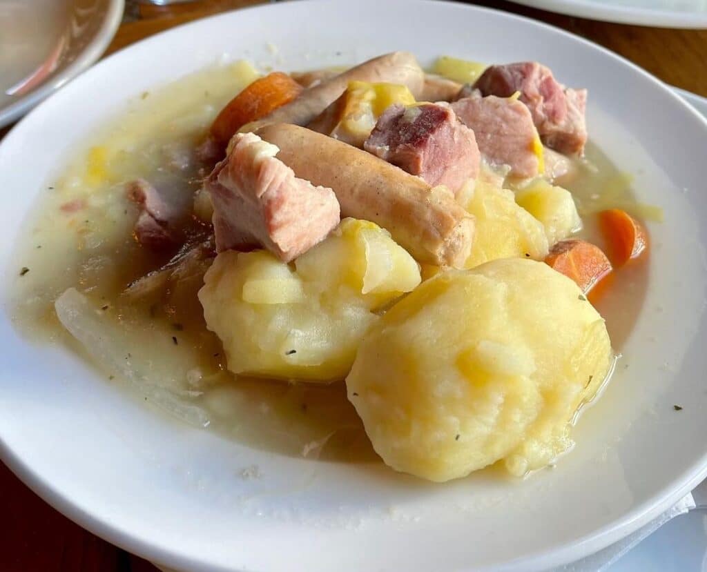 A white bowl filled with a traditional Dublin coddle, showing pale boiled sausages, pieces of back bacon, large cooked potatoes, and sliced carrots in a clear broth.