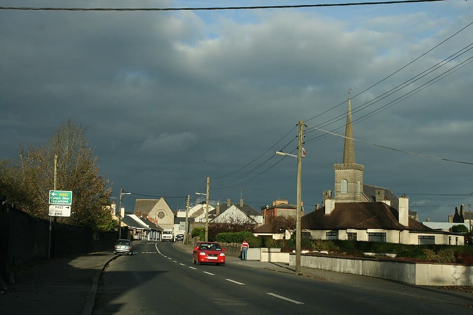 A scenic road view entering Mountmellick, County Laois, featuring a prominent church steeple against a dark, cloudy sky and a red car driving through the town.