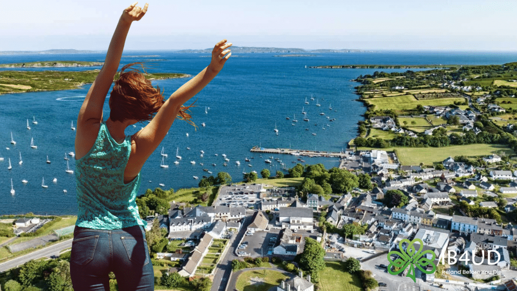 A woman with her arms raised stands on a high vantage point overlooking the scenic coastal town of Schull, West Cork, with its harbour filled with sailboats.