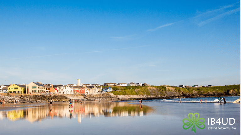 A panoramic view of Lahinch beach in Co. Clare, showing visitors on the wet sand reflecting colourful seafront buildings and a clear blue sky.